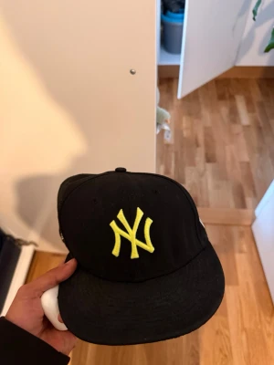 Svart New Era Yankees keps - Säljer en svart New Era keps med gul New York Yankees-logga framtill. Kepsen har platt skärm och broderade loggor på sidan och baksidan. Skärmen har gul detalj undertill. Perfekt för dig som vill ha en sportig och trendig look.