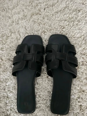 Trendiga från hm helt nya - Säljer ett par stilrena svarta slip-in sandaler med breda remmar och fyrkantig tå. Sandalerna har en modern design med dubbla remmar i konstläder och öppen häl. Perfekta för en trendig och avslappnad look.