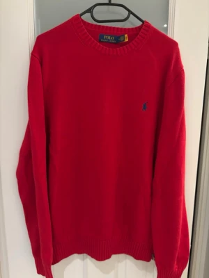 Röd stickad tröja från Polo Ralph Lauren - Klassisk röd stickad tröja från Polo Ralph Lauren med rund halsringning och blå broderad logga på bröstet. Tröjan har ribbstickade muddar vid ärmslut och nederkant. Perfekt för dig som gillar stilrena och tidlösa plagg.