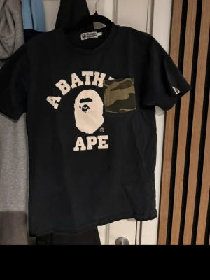 Svart BAPE t-shirt med camoficka - Säljer en svart t-shirt från A Bathing Ape med stor vit logga och ikoniskt apansikte på bröstet. T-shirten har en unik bröstficka i camouflage-mönster som sticker ut. Klassisk passform och rund hals, perfekt för streetwear-stilen.