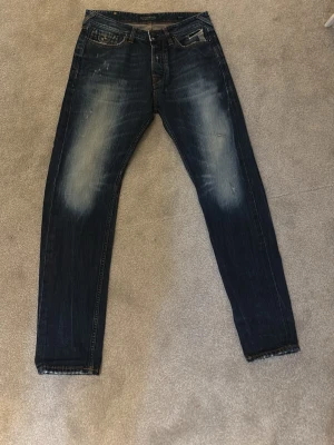 Mörkblå Dean jeans från Scotch & Soda - Snygga mörkblå Dean jeans från Scotch & Soda med slitna detaljer och ljusa partier på låren. Klassisk femficksmodell med raka ben och orange kontrastsömmar. Perfekta för dig som gillar en avslappnad men trendig look.