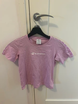 Rosa Champion t-shirt med logga - Snygg rosa t-shirt från Champion med vit logga på bröstet och dekorativa band med Champion-mönster längs axlarna. Klassisk rund halsringning och korta ärmar. Perfekt för en avslappnad och sportig stil.