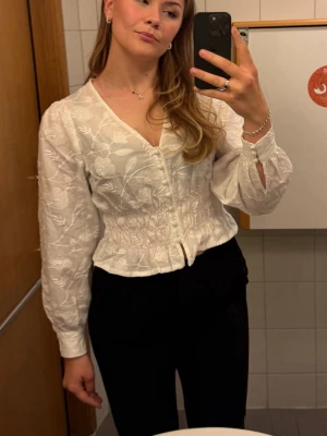 Vit blus med broderade blommor - Söt vit blus från Holly & Whyte med broderade blommor, v-ringning och knappar framtill. Blusen har lång ärm med resår i midjan och vid ärmslut. Materialet är lätt och ger en romantisk känsla. Perfekt till jeans eller kjol.
