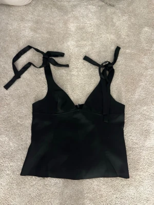 Svart linnetopp från Gina Tricot - Snygg svart linnetopp från Gina Tricot med breda axelband som knyts i rosetter. Har i vanliga fall S i vanlig storlek. Har sytt upp banden vid axlarna men är enkelt att bara sprätta ut ifall det skulle behövas! Använd 1 gång