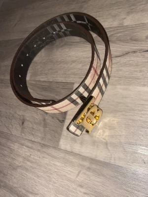 Rutig Burberry-bälte med guldspänne - Snyggt bälte från Burberry med klassiskt rutigt mönster i beige, svart och rött. Bältet har ett ikoniskt guldigt BB-spänne som verkligen sticker ut. Perfekt accessoar för att lyfta vilken outfit som helst.