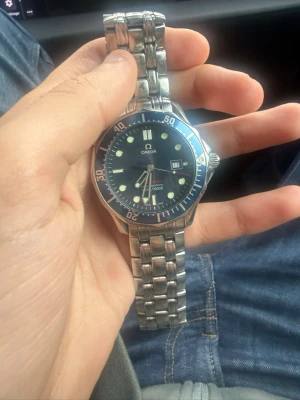 Omega Seamaster blå klocka i stål - Säljer en Omega Seamaster klocka med blå urtavla och datumvisning. Klockan har en robust länk i rostfritt stål och vridbar bezel. Klassisk dykardesign med tydliga lysande markeringar och visare. Perfekt för dig som gillar stilrena accessoarer.