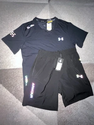 Under Armour Set - Snyggt svart träningsset från Under Armour med t-shirt och shorts. Båda plaggen har logga och text i regnbågsfärger, samt elastisk midja med dragsko på shortsen. Perfekt för gymmet eller löprundan.