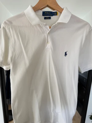 Vit slim fit pikétröja från Polo Ralph Lauren - Vit pikétröja från Polo Ralph Lauren i slim fit stretch mesh. Klassisk krage, knappar framtill och ikonisk broderad logga på bröstet. Perfekt för dig som gillar en clean och stilren look.