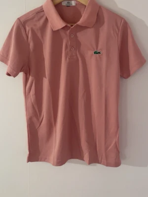 Rosa pikétröja från Lacoste - Klassisk rosa pikétröja från Lacoste med korta ärmar och krage. Tröjan har knappar framtill och den ikoniska gröna krokodilloggan på bröstet. Tillverkad i mjuk bomull som känns skön mot huden. Perfekt för en avslappnad men ändå stilsäker look.