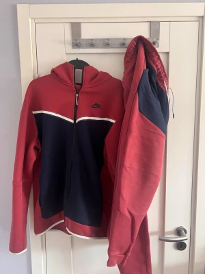 Nike tech röd och marinblå tracksuit - Snygg hoodie från Nike i rött och marinblått med dragkedja framtill och vit detalj över bröstet. Klassisk Nike-logga på vänster sida. Huva med dragsko och ribbade muddar. Perfekt för en sportig och avslappnad stil. SE BILDER FÖR DEFEKT
