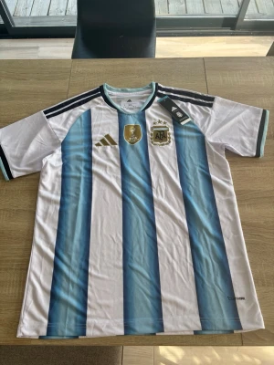 Argentina matchtröja Adidas blåvit - Säljer en officiell Argentina matchtröja från Adidas med klassiska blå och vita vertikala ränder. Tröjan har svarta detaljer på axlarna, guldiga logotyper och broderade märken på bröstet. Tillverkad i lätt och ventilerande polyester, perfekt för fotboll.