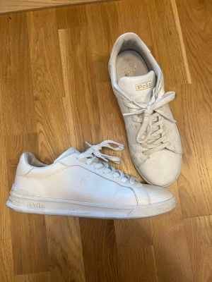 Vita Polo Ralph Lauren sneakers - Snygga vita sneakers från Polo Ralph Lauren i klassisk low-top modell. Skorna är tillverkade i 100% läder med textil- och läderfoder samt gummisula. Diskret Polo-logga på sidan och hälen. Perfekta för en clean och stilren look.