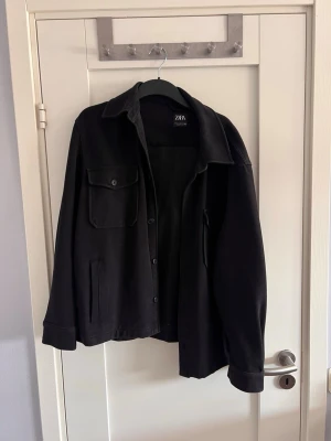 Svart overshirt från Zara - Stilren svart overshirt från Zara med klassisk krage, knappstängning och två fickor framtill, varav en med lock. Overshirten har en rak passform och är tillverkad i ett mjukt tyg som ger en avslappnad look.
