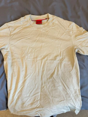  HUGO - Tshirt  - Stilren beige t-shirt från HUGO med diskret broderad logga på bröstet. Klassisk rund hals och korta ärmar. Tillverkad i mjuk bomull för en bekväm känsla och enkel att matcha med olika outfits.