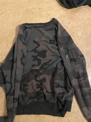 Camouflage sweatshirt med ficka - Säljer en mörkgrön och svart camouflage sweatshirt från Alpha Industries. Tröjan har en unik ficka med dragkedja på ena ärmen och ribbade muddar vid ärmslut och nederkant. Perfekt för dig som gillar streetwear och vill sticka ut lite extra.