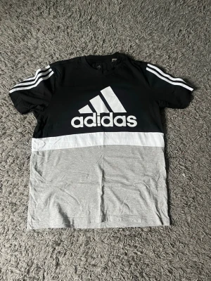 Svart och grå Adidas t-shirt - Snygg Adidas t-shirt i svart, vitt och grått med stor logga framtill och klassiska vita ränder på axlarna. T-shirten har rund hals och korta ärmar. Tillverkad i mjuk bomull, perfekt för en sportig och avslappnad stil.