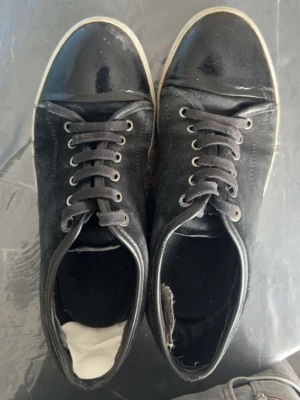 Lanvins  - Säljer ett par svarta sneakers med klassisk snörning och snygg lackad tå. Skorna har en vit sula och är tillverkade i textil och syntetmaterial. Perfekta för dig som gillar en stilren och enkel look.