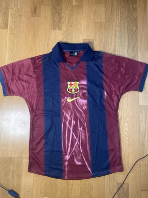 Travis Scott X Nike X Barcelona dri fit shirt - Säljer en klassisk FC Barcelona matchtröja från Nike i vinrött och marinblått med klubbens emblem på bröstet och gul Nike-logga. Tröjan har korta ärmar, krage och är tillverkad i ett lätt, ventilerande material perfekt för fotbollsträning. Rekommenderar PostNord för snabbare leverans
