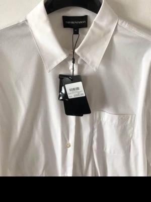 Vit skjorta från Emporio Armani - Klassisk vit skjorta från Emporio Armani med normal passform. Skjortan har lång ärm, bröstficka med diskret logga och snygga knappar med märkets namn. Tillverkad i mjuk bomull för en clean och stilren look.