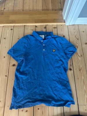 Blå pikétröja från Lyle & Scott - Snygg blå pikétröja från Lyle & Scott med klassisk krage och knappar framtill. Tröjan har det ikoniska gula logomärket på bröstet och är tillverkad i mjuk bomull. Perfekt för en avslappnad och stilren look.