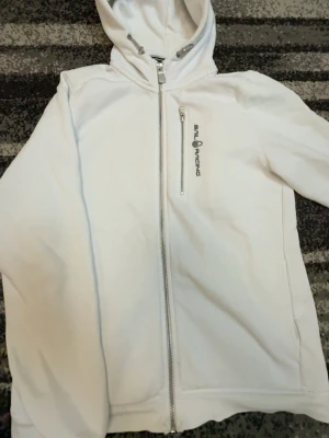 Vit hoodie från Sail Racing - Snygg vit hoodie från Sail Racing med dragkedja framtill och en extra dragkedjeförsedd bröstficka. Hoodien har justerbar huva med snören och logga broderad på bröstet. Perfekt för en clean och sportig stil.