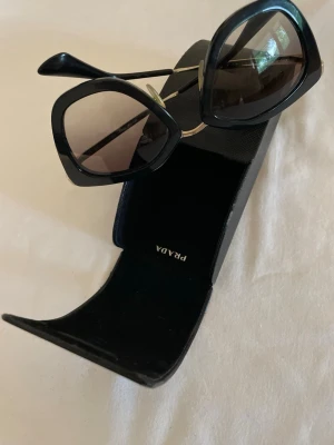 Svarta solglasögon från Prada - Snygga svarta solglasögon från Prada med stora, fyrkantiga bågar och mörka glas. Klassisk och trendig design med en lyxig känsla. Kommer med originalfodral. Perfekta för att höja din stil till nästa nivå.