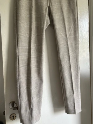 Beige rutiga kostymbyxor - Snygga beige kostymbyxor med diskret rutmönster. Byxorna har raka ben och pressveck framtill för en clean look. Perfekta till en dressad outfit och passar till både skjorta och t-shirt. Materialet känns mjukt och har en klassisk känsla.