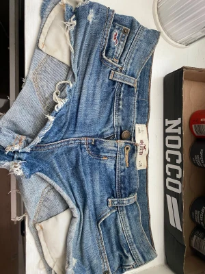 Blå jeansshorts från Hollister - Säljer ett par klassiska blå jeansshorts från Hollister med fransiga kanter och slitna detaljer. Shortsen har fem fickor, bälteshällor och knappgylf. Perfekta för varma dagar och ger en avslappnad vibe.