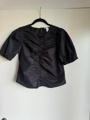 Svart puffärmad blus med rynk - Stilren svart blus med puffärmar och rynkdetaljer framtill. Blusen har rund halsringning och dragkedja i ryggen. Tillverkad i ett glansigt, vävt material som ger en cool look. Perfekt för dig som gillar trendiga detaljer och enkel design.