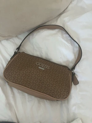 Beige axelremsväska från Guess - Snygg beige handväska från Guess med klassiskt logomönster över hela väskan och silverfärgad logga framtill. Väskan har en kort axelrem och dragkedja upptill. Perfekt storlek för det viktigaste och ger en trendig touch till din outfit. Den är använd men fortfarande i väldigt fint skick, finns några slitningar på undersidan av väskan.