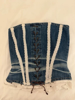 Jeanskorsett med spetsdetaljer - Säljer en snygg korsett i blå denim med vita spetsband och snörning framtill. Mått finns i bilderna💙