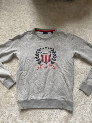 Grå sweatshirt från GANT med emblem - Snygg grå sweatshirt från GANT med broderat emblem framtill i marinblått, rött och vitt. Tröjan har rund halsringning, ribbade muddar och klassisk collegekänsla. Perfekt för en avslappnad och sportig stil.