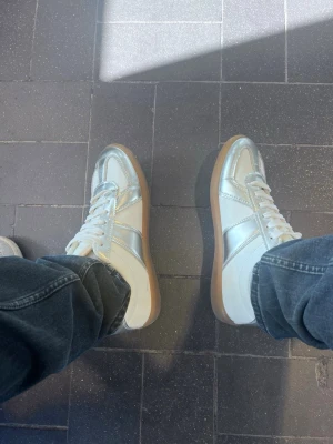 Vita och silver sneakers med gummisula - Säljer ett par stilrena sneakers i vitt och silver med klassisk låg profil och gummisula. Skorna har vita snören och paneler i metallic-silver som ger en modern touch. Perfekta för dig som vill sticka ut med en clean look. Materialet är en mix av läder och textil.