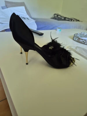 Svarta pumps med fjädrar och gulddetalj - Unika svarta pumps från Zara Woman Collection med öppen tå, smal guldfärgad klack och dekorativa svarta fjädrar framtill. Skorna har en elegant siluett och materialet ser ut att vara satinliknande tyg. Perfekta för dig som vill sticka ut.