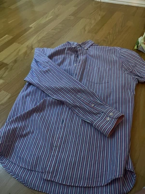 Randig skjorta från MYLA, blå/rosa - Säljer en långärmad skjorta från MYLA i storlek L. Skjortan har smala blå, rosa och vita ränder, klassisk krage och knäppning framtill. Tillverkad i ett mjukt bomullstyg som känns skönt mot huden. Perfekt för dig som gillar stilrena och färgglada plagg.