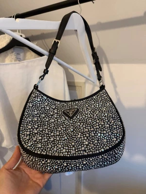 Prada svart axelväska med strass - Supersnygg svart axelväska från Prada täckt med glittrande strass-stenar. Väskan har en klassisk halvmåneform och justerbart axelband i svart läder. Perfekt för dig som vill sticka ut och addera lite extra bling till din outfit.