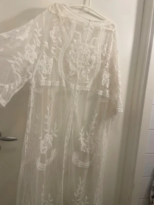 Vit spetskimono strandplagg onesize - Säljer en superfin vit kimono i spets med blommiga broderier, perfekt som strandplagg. Lång modell med vida ärmar och transparent material. Tillverkad i 50% bomull och 50% polyamid. Ger en romantisk och bohemisk vibe till din strandlook.