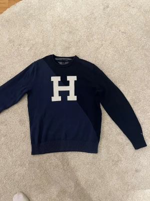 Mörkblå stickad tröja Tommy Hilfiger - Mörkblå stickad tröja från Tommy Hilfiger med stor vit H på bröstet. Tröjan har rund halsringning, ribbade muddar och är tillverkad i mjukt material. Perfekt för dig som gillar klassisk college-stil.