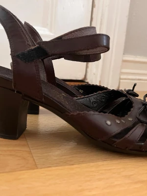 Bruna sandaler med klack och remmar - Säljer ett par mörkbruna sandaler med klack och justerbara ankelremmar. Sandalerna har öppen tå, dekorativa nitar och flätade detaljer framtill. Tillverkade i läderliknande material och passar perfekt till sommarens outfits. Vintage!