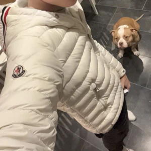 Moncler dunjacka❄️ - Säljer en snygg vit dunjacka från Moncler med huva och dragkedja. Jackan har ett stilrent märke på ärmen och ränder i rött, vitt och blått på huvan. 