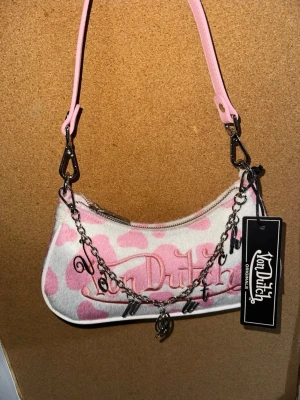 Rosa och vit Von Dutch handväska - Trendig handväska från Von Dutch i rosa och vitt med hjärtmönster. Väskan har en rosa axelrem, silverfärgad kedja med bokstavsberlocker och broderad logga framtill. Materialet är mjukt tyg och väskan stängs med dragkedja. Perfekt accessoar för en cool streetstyle-look.