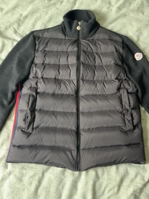 Moncler Cardigan - Snygg svart pufferjacka från Moncler i storlek S med stickade ärmar och hög krage. Jackan har två fickor framtill med knappstängning och dragkedja i mitten. Moncler-logga på vänster ärm. Perfekt för både vårdagar samt kalla vinterdagar. Finns i Stockholm går även att fraktas.