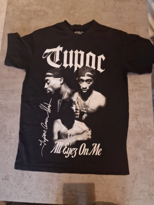 Svart Tupac t-shirt Pull&Bear - Säljer en svart t-shirt från Pull&Bear med stort Tupac-tryck och texten 'All Eyez On Me' framtill. T-shirten har klassisk passform och är tillverkad i mjuk bomull. Perfekt för dig som gillar streetwear och ikoniska musikmotiv. Står small men passar säkert som medium
