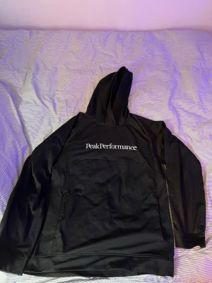 Svart hoodie från Peak Performance - Snygg svart hoodie från Peak Performance med stor vit logga på bröstet. Klassisk huva och känguruficka framtill. Tillverkad i mjukt och bekvämt material som passar perfekt till en avslappnad stil.