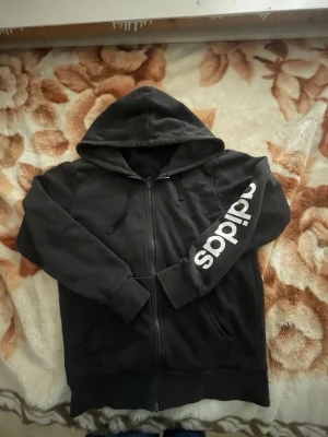 Svart hoodie med dragkedja från Adidas - Svart hoodie från Adidas med dragkedja framtill och stor huva. Ärmen har en stor vit Adidas-logga tryckt längs sidan. Hoodien har ribbade muddar och fickor på framsidan. Perfekt för en avslappnad streetwear-look.