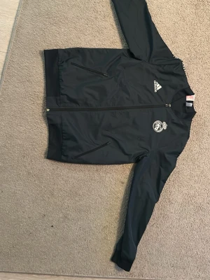 Svart Adidas Real Madrid jacka - Snygg svart träningsjacka från Adidas med Real Madrid-logga på bröstet och vita Adidas-detaljer. Jackan har dragkedja framtill, två fickor med dragkedja och ribbade muddar vid ärmslut och nederkant. Perfekt för träning eller till vardags.