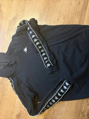 Svart Kappa hoodie med logoband - Svart hoodie från Kappa med klassiska vita logoband längs ärmarna och broderad logga på bröstet. Hoodien har huva med dragsko och en stor magficka. Tillverkad i mjukt material som känns skönt mot huden. 