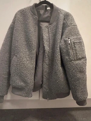 Grå teddyjacka med dragkedja - Säljer en oversized grå teddyjacka med mjukt, fluffigt material och ribbade muddar. Jackan har en dragkedja framtill, ficka med dragkedja på ärmen och bomberjacke-look. Perfekt för kalla dagar och riktigt snygg till streetwear-stil.