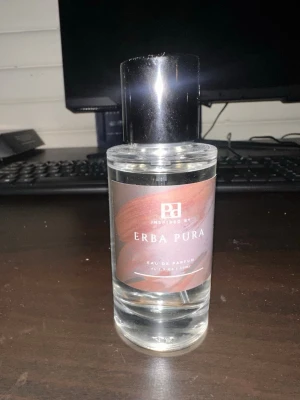Erba Pura Eau de Parfum - En stilren parfymflaska med svart lock och transparent glas. Etiketten är brun med vit text och har en modern design. Doften heter Erba Pura och är Eau de Parfum. Perfekt för dig som gillar exklusiva och fräscha dofter. Endast testad