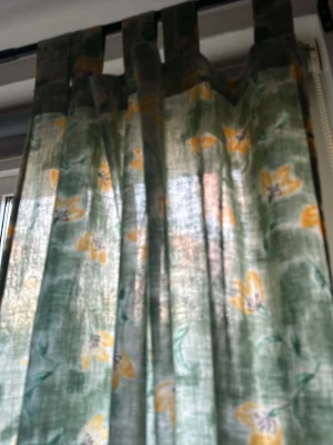 Gröna gardiner med gula blommor - Säljer ett par gardiner i tunn grön textil med gula blommor och gröna blad. Gardinerna har kanal upptill för enkel upphängning och ett lätt transparent tyg som släpper in ljus. Perfekt för att ge rummet en fräsch och somrig känsla.